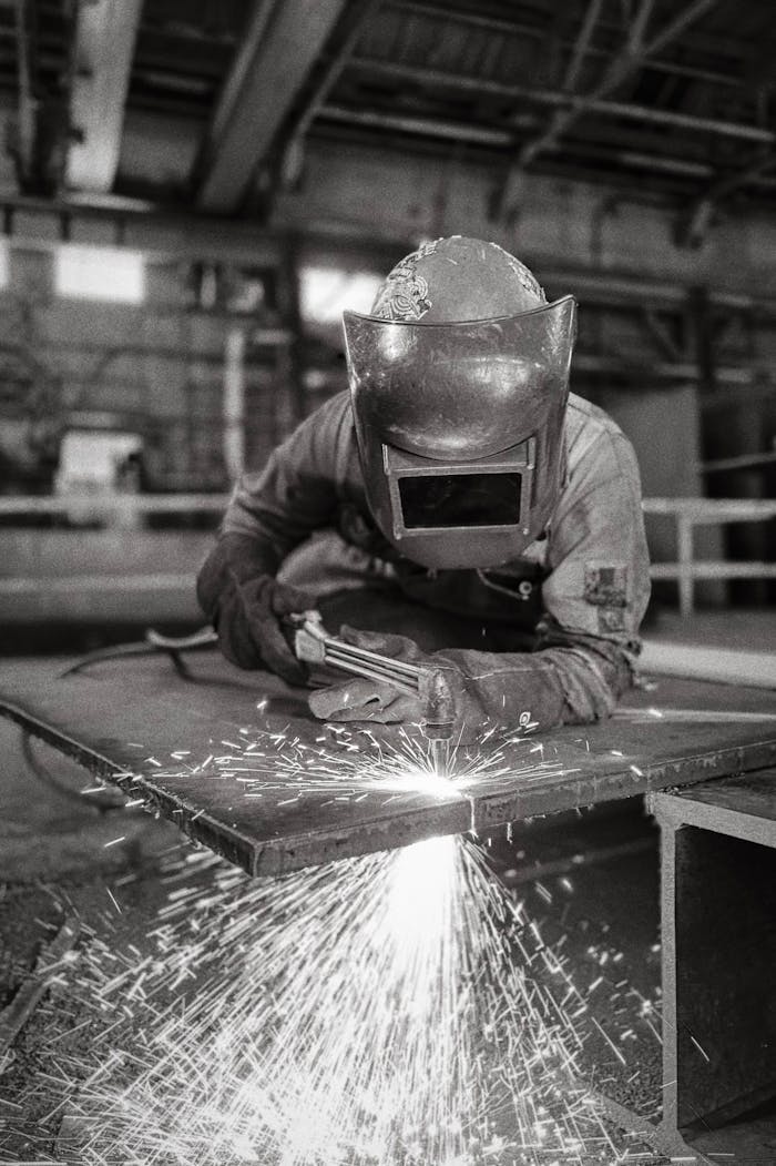 A welder wearing protective gear sparks metal in an industrial factory setting.